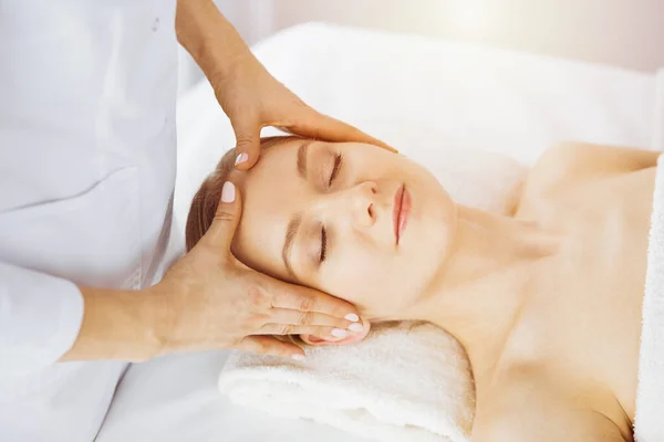 Hermosa mujer caucásica disfrutando de masaje facial con los ojos cerrados en el salón de spa soleado. Tratamiento relajante en medicina y concepto de belleza — Foto de Stock