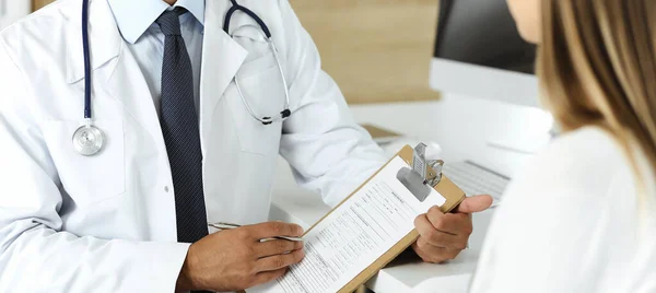 Docteur et patient discutant des résultats de l'examen médical assis au bureau de la clinique, gros plan. Médecin masculin utilisant presse-papiers pour remplir les antécédents médicamenteux de la jeune femme — Photo