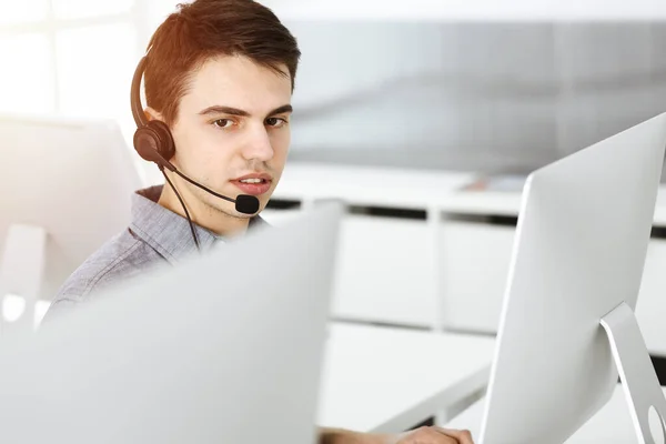 Casual vestido jovem usando fone de ouvido e computador enquanto conversa com clientes on-line no escritório ensolarado. Call center, conceito de negócio — Fotografia de Stock