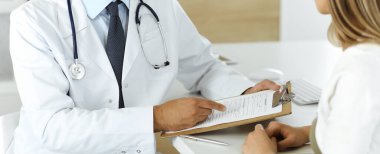 Doktor ve hasta klinikteki masada otururken tıbbi muayene sonuçlarını tartışıyorlar, yakın plan. Genç bir kadının ilaç geçmişini doldurmak için pano kullanan bir erkek doktor.