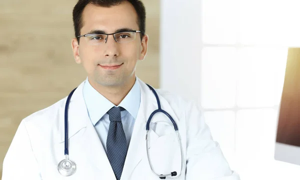 Hombre-médico sentado en el escritorio en su lugar de trabajo y sonriendo. Servicio médico perfecto en clínica soleada —  Fotos de Stock