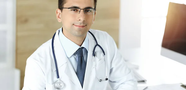 Man-dokter zit aan het bureau op zijn werkplek en glimlacht. Perfecte medische dienst in zonnige kliniek — Stockfoto