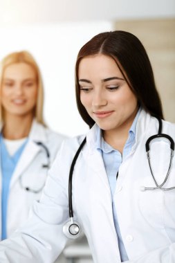 Esmer kadın doktor, güneşli klinikte pano kullanırken ilaç geçmişi ve sınav sonuçlarını kontrol ediyor.