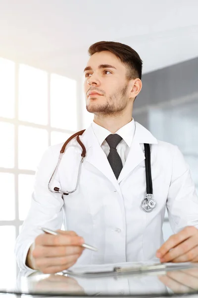 Γιατρός άνθρωπος συμπληρώνοντας έντυπο ιστορικό φαρμάκων, ενώ κάθεται στο γυάλινο γραφείο στην ηλιόλουστη κλινική. Έννοια ιατρικής — Φωτογραφία Αρχείου