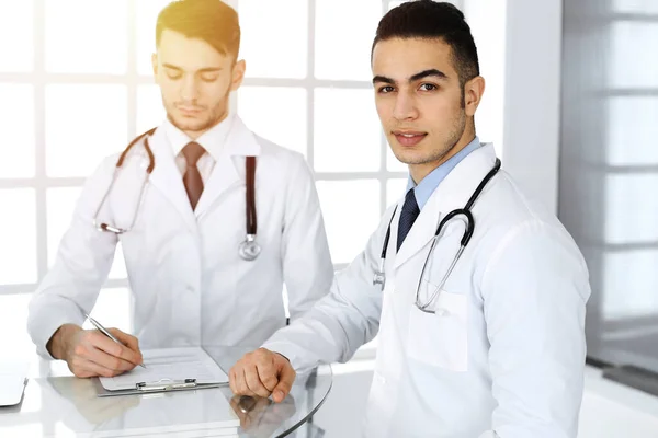 Άραβας γιατρός κάθεται στο γυάλινο γραφείο με καυκάσιο συνάδελφο στο ιατρικό γραφείο ή ηλιόλουστη κλινική. Διαφορετική ομάδα γιατρών, ιατρική έννοια — Φωτογραφία Αρχείου