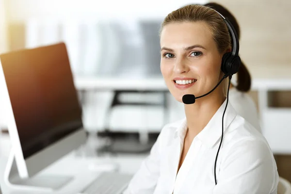 Mujer rubia representante de servicio al cliente está consultando a los clientes en línea utilizando auriculares en la oficina soleada. Concepto de Call center — Foto de Stock