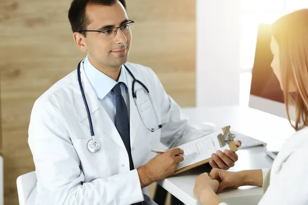 Läkare och patient diskuterar medicinska undersökningsresultat medan du sitter vid skrivbordet i solig klinik — Stockfoto