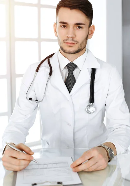 Médecin homme remplir les dossiers de l'histoire des médicaments formulaire alors qu'il était assis au bureau en verre dans une clinique ensoleillée. Concept de médecine — Photo