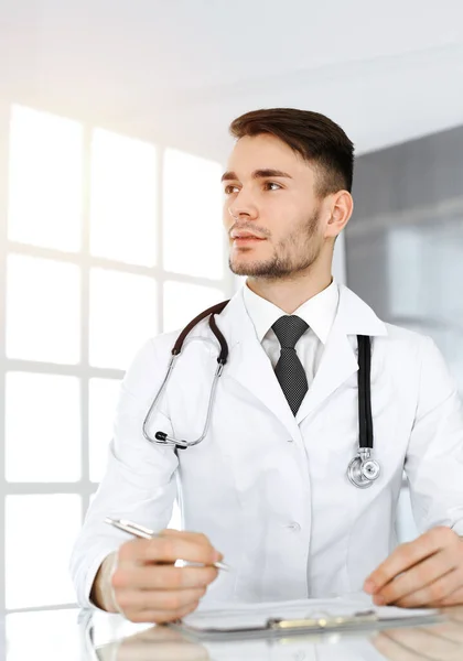 Γιατρός άνθρωπος συμπληρώνοντας έντυπο ιστορικό φαρμάκων, ενώ κάθεται στο γυάλινο γραφείο στην ηλιόλουστη κλινική. Έννοια ιατρικής — Φωτογραφία Αρχείου