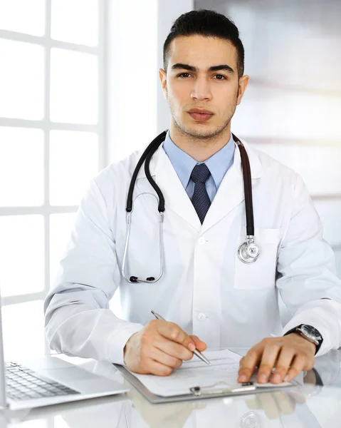 Arabischer Arzt benutzt Laptop-Computer, während er Medikamentenanamnese aus sonniger Klinik auffüllt. Medizinkonzept — Stockfoto