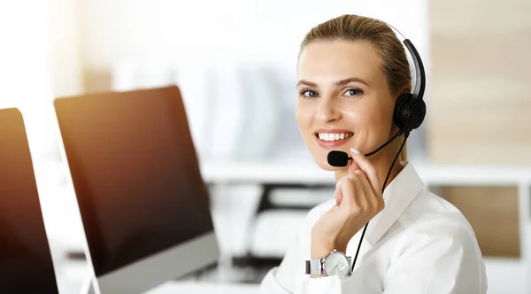 Blond female customer service representative is consulting clients online using headset in sunny office. Call center concept