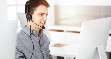 Güneşli ofiste müşterilerle konuşurken kulaklık ve bilgisayar kullanan sıradan giyinmiş genç bir adam. Çağrı merkezi, iş konsepti