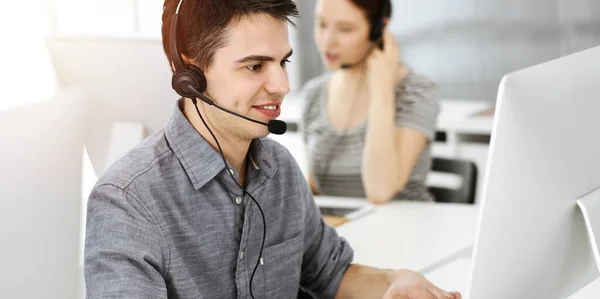 Hombre joven vestido casual usando auriculares y computadora mientras habla con los clientes en línea en la oficina soleada. Call center, concepto de negocio — Foto de Stock