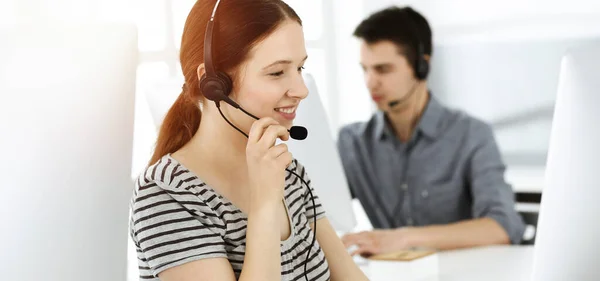 Casual dressed young woman using headset and computer while talking with customers online. Group of operators at work in sunny office. Call center — Stock Photo, Image