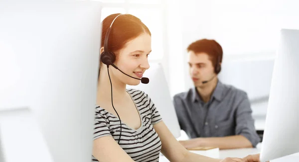 Mujer joven vestida casual usando auriculares y computadora mientras habla con los clientes en línea. Grupo de operadores en el trabajo en la oficina soleada. Centro de llamadas — Foto de Stock