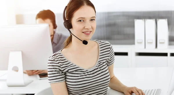 Casual dressed young woman using headset and computer while talking with customers online. Group of operators at work in sunny office. Call center