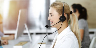 Sarışın iş kadını iletişim kurmak ve güneşli ofisteki insanlara danışmak için kulaklık kullanıyor. Çağrı merkezi