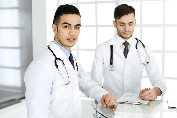 Άραβας ιατρός κάθεται στο γυάλινο γραφείο με καυκάσιο συνάδελφο στο ιατρικό γραφείο ή κλινική. Διαφορετική ομάδα ιατρών, ιατρική και υγειονομική περίθαλψη έννοια — Φωτογραφία Αρχείου