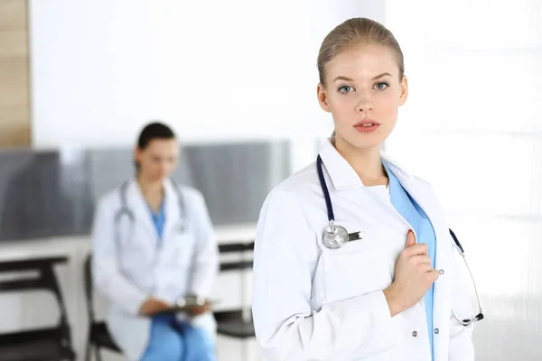 Mujer - médico de pie en la clínica con su colega en el fondo. Médico trabajando. Concepto de medicina y salud —  Fotos de Stock