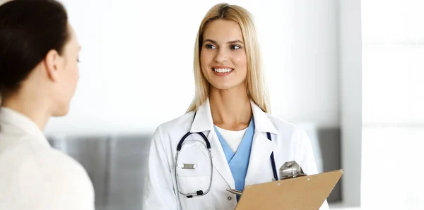 Woman-doctor at work in hospital is happy to consult female patient. Blonde physician checks medical history record and exam results while using clipboard. Medicine concept