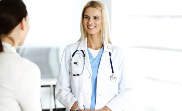 Mujer-médico en el trabajo en el hospital está feliz de comunicarse con la paciente femenina. Médico rubio revisa historial médico y resultados del examen. Concepto de medicina — Foto de Stock