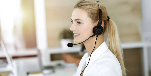 Mujer rubia de negocios que usa auriculares para comunicarse y consultar a la gente en la oficina soleada. Centro de llamadas — Foto de Stock