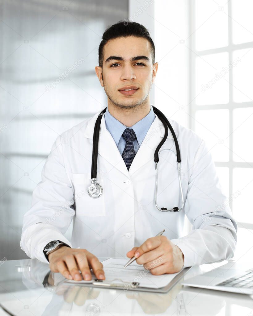 Arab doctor man is using laptop computer while filling up medication history records form at the glass desk in r clinic. Medicine concept