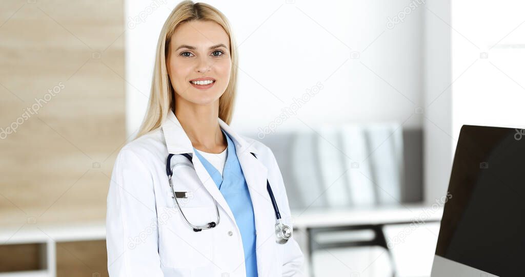 Blond female physician is standing at her workplace near desktop computer. Woman-doctor is excited and happy of her profession. Medicine concept