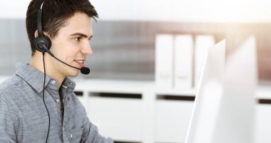 Güneşli ofiste müşterilerle konuşurken kulaklık ve bilgisayar kullanan sıradan giyinmiş genç bir adam. Çağrı merkezi, iş konsepti