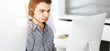 Güneşli ofiste müşterilerle konuşurken kulaklık ve bilgisayar kullanan sıradan giyinmiş genç bir adam. Çağrı merkezi, iş konsepti