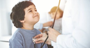Güneşli klinikte bir çocuk hastayı steteskopla muayene eden doktor-kadın. Tatlı Arap çocuk ve kardeşi doktor randevusundalar.