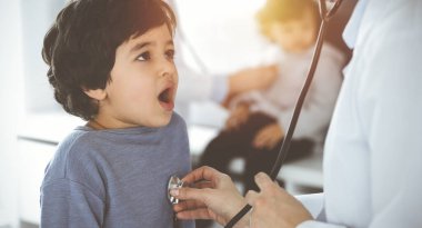 Güneşli klinikte bir çocuk hastayı steteskopla muayene eden doktor-kadın. Tatlı Arap çocuk ve kardeşi doktor randevusundalar.