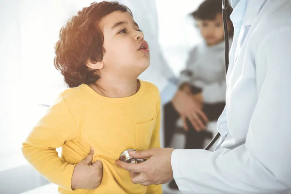 Ženský lékař vyšetřuje dětského pacienta stetoskopem na slunné klinice. Roztomilý arabský chlapec na návštěvě lékaře — Stock fotografie