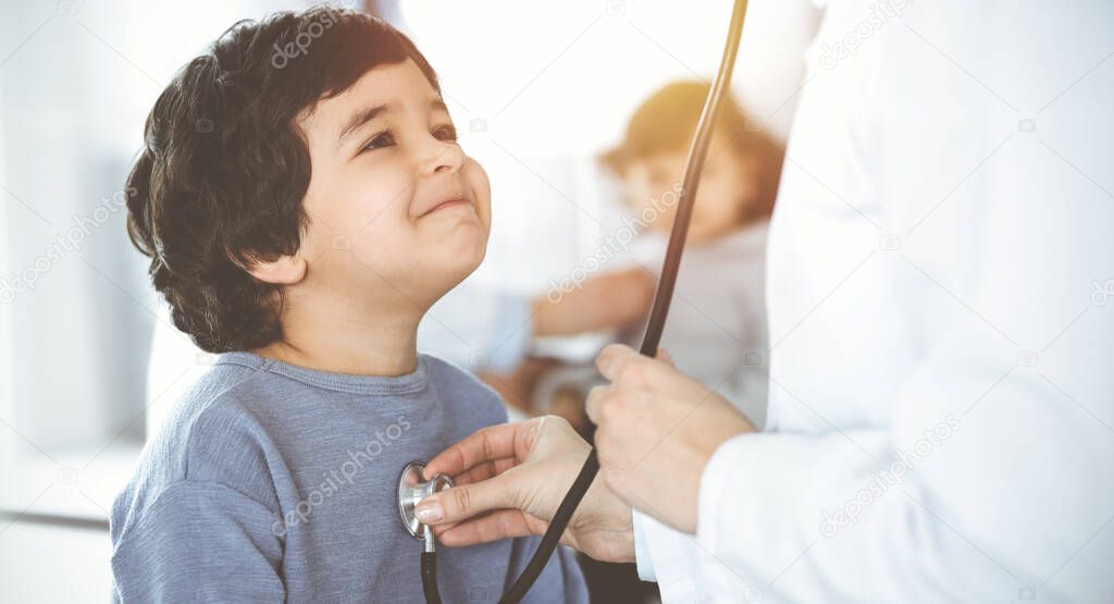 Doctor-woman examining a child patient by stethoscope in sunny clinik. Cute arab boy and his brother at physician appointment