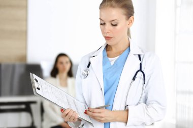 Kadın - doktor hastanın arka planında klinikte tıbbi geçmiş kayıtlarını doldurmak için pano kullanıyor. Tıp konsepti