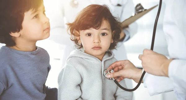 Médico-mujer examinando a un niño paciente por estetoscopio en Sunny Clinik. Lindo chico árabe y su hermano en la cita médica —  Fotos de Stock