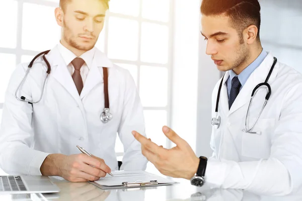 Médico árabe sentado en el escritorio de cristal con colega caucásico en el consultorio médico o clínica soleada. Equipo de médicos diversos, concepto de medicina — Foto de Stock