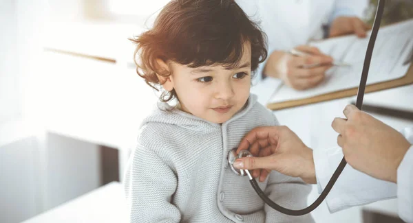 Mujer-médico examinando a un niño paciente por estetoscopio en el soleado Clinik. Lindo niño árabe en la cita con el médico —  Fotos de Stock