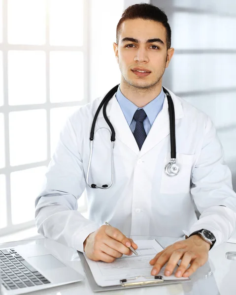 Arabische arts man is met behulp van laptop computer tijdens het vullen van medicatie geschiedenis records formulier zonnige kliniek. Geneesmiddelenconcept — Stockfoto