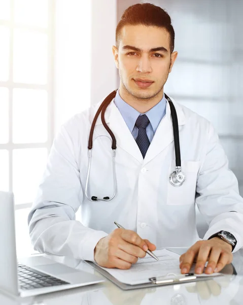 Arabo medico uomo sta utilizzando il computer portatile durante la compilazione record di storia di farmaci forma clinica soleggiata. Concetto di medicina — Foto Stock