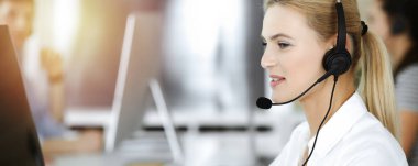 Sarışın iş kadını iletişim kurmak ve güneşli ofisteki insanlara danışmak için kulaklık kullanıyor. Çağrı merkezi