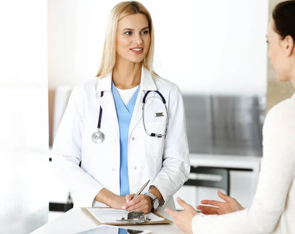 Woman-doctor at work in sunny hospital is happy to consult female patient. Blonde physician checks medical history record and exam results while using clipboard