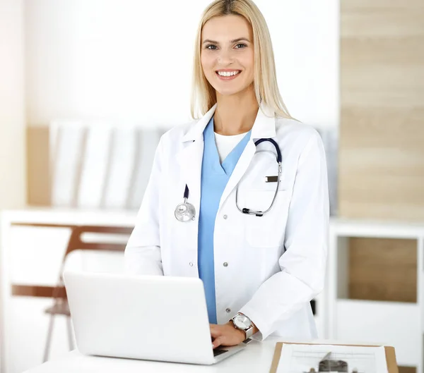 Woman-doctor controls medication history records and exam results while using laptop computer in sunny hospital. Medicine and healthcare concept — Stock Photo, Image