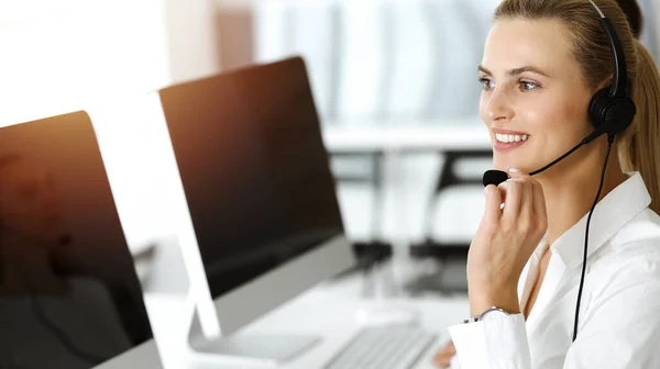 ブロンドの女性のカスタマーサービス担当者は、日当たりの良いオフィスでヘッドセットを使用してオンラインでクライアントに相談しています。コールセンターのコンセプト — ストック写真