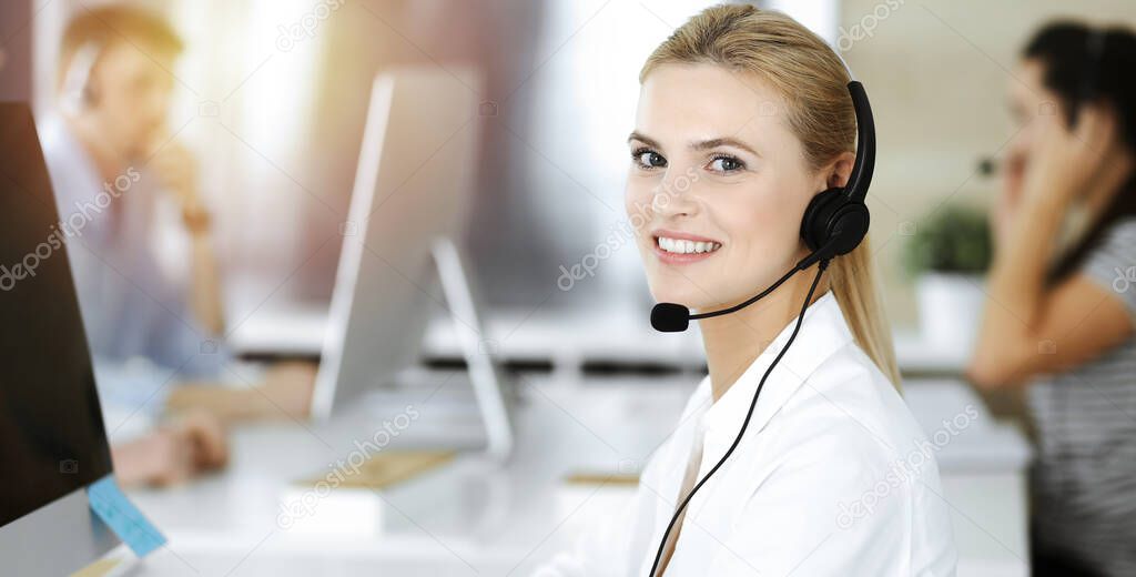 Blond business woman using headset for communication and consulting people at sunny office. Call center