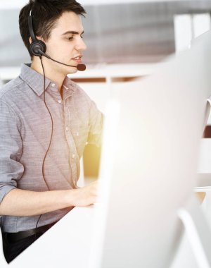 Güneşli ofiste müşterilerle konuşurken kulaklık ve bilgisayar kullanan sıradan giyinmiş genç bir adam. Çağrı merkezi, iş konsepti