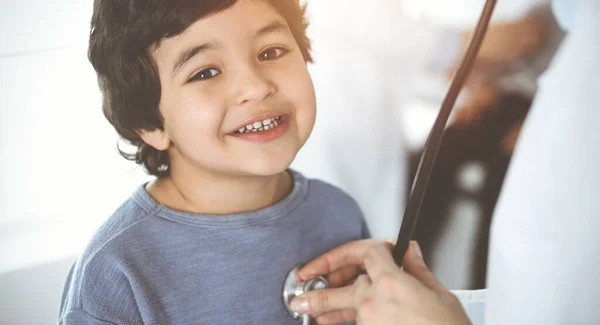 Doctor-woman examining a child patient by stethoscope in sunny clinik. Cute arab boy and his brother at physician appointment — Stock Photo, Image