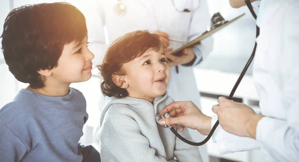 Médico-mujer examinando a un niño paciente por estetoscopio en Sunny Clinik. Lindo chico árabe y su hermano en la cita médica —  Fotos de Stock