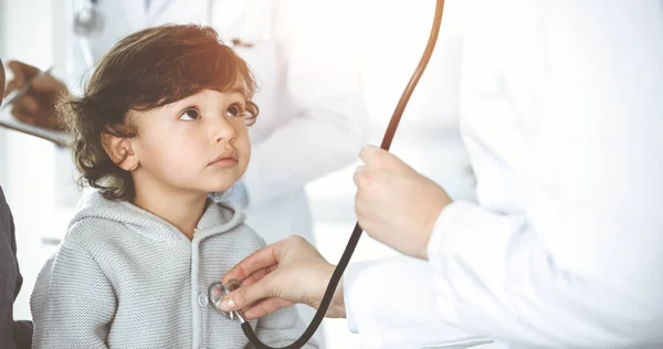 Médico-mujer examinando a un niño paciente por estetoscopio en Sunny Clinik. Lindo chico árabe y su hermano en la cita médica —  Fotos de Stock