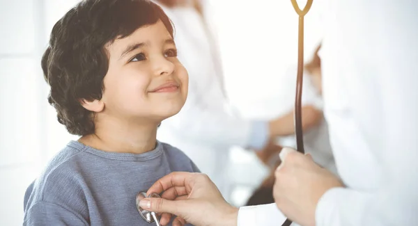 Médico-mulher examinando uma criança paciente por estetoscópio em clinik ensolarado. bonito árabe menino e seu irmão no médico nomeação — Fotografia de Stock